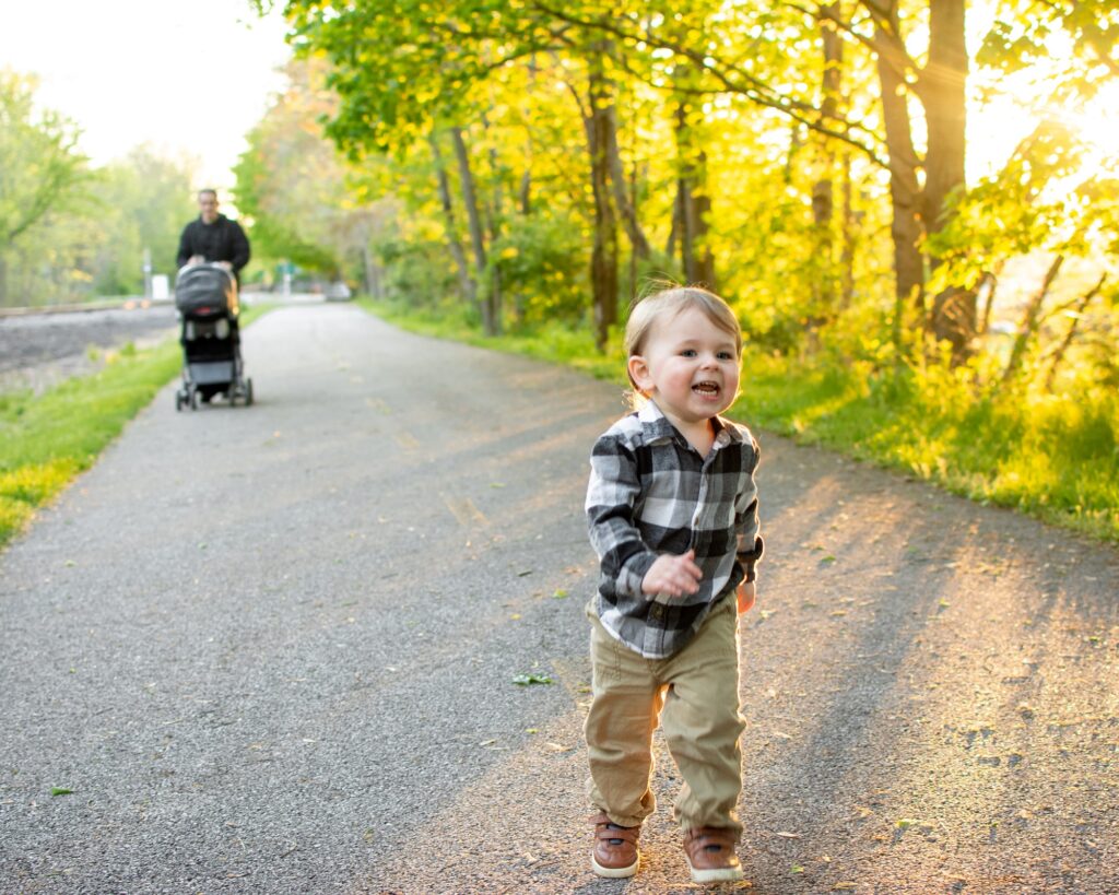 Young toddler running on a paved path, while his dad pushes a stroller behind him.